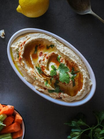 Lebanese hummus serves on a plate with a bowl of carrot and celery sticks.