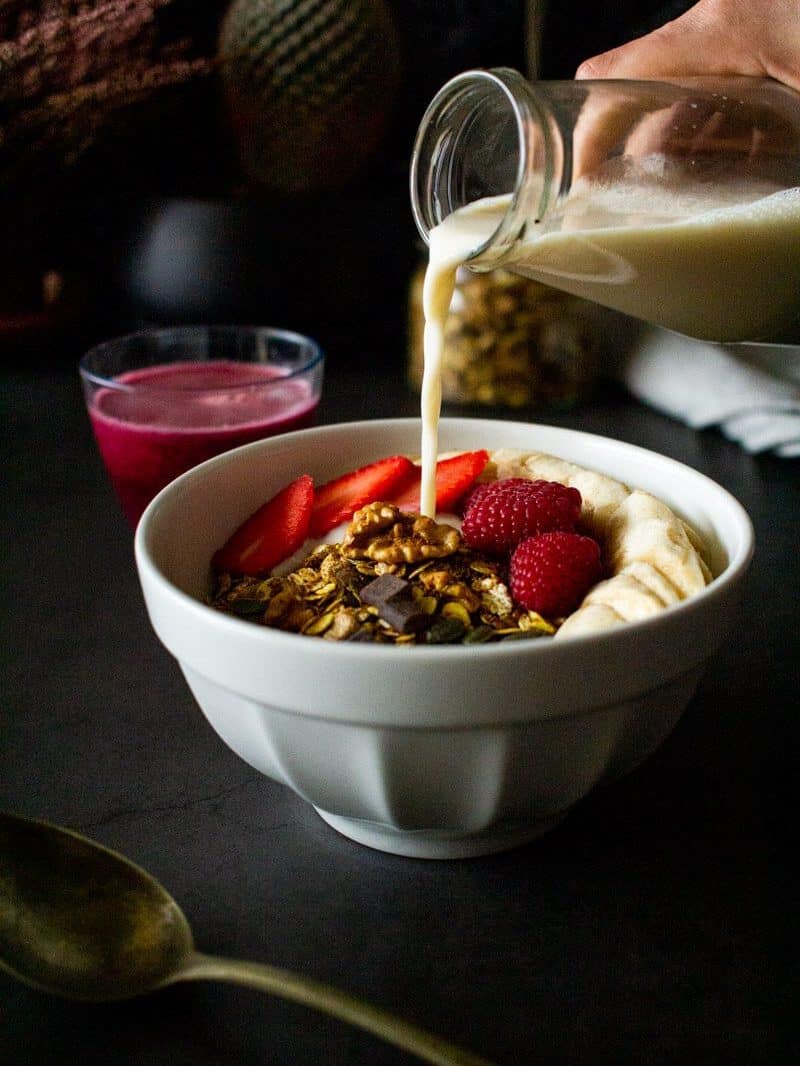 pouring plant milk over a bowl with our gluten-free granola recipe and strawberries