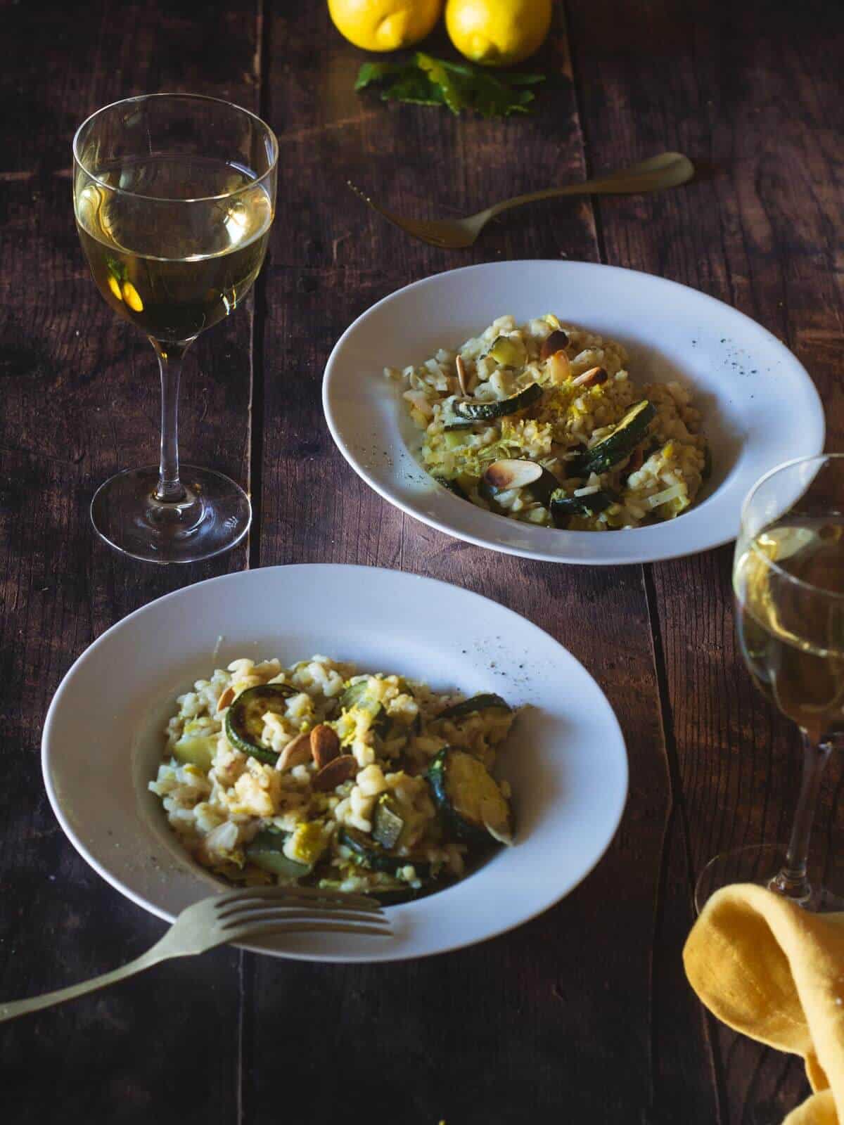 dos platos de risotto de verduras sobre mesa de madera con dos copas de vino blanco.
