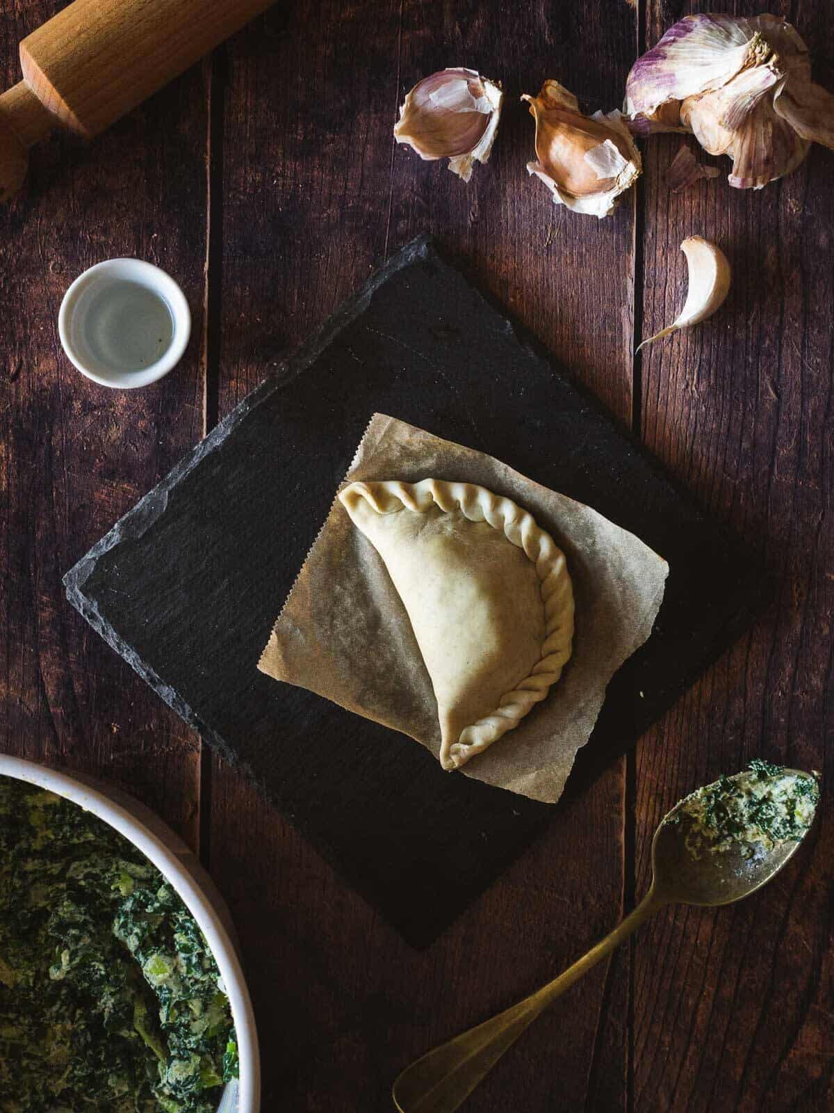 uncooked spinach empanada closed with folding technique over parchment paper with little bowl of water to get fingers wet