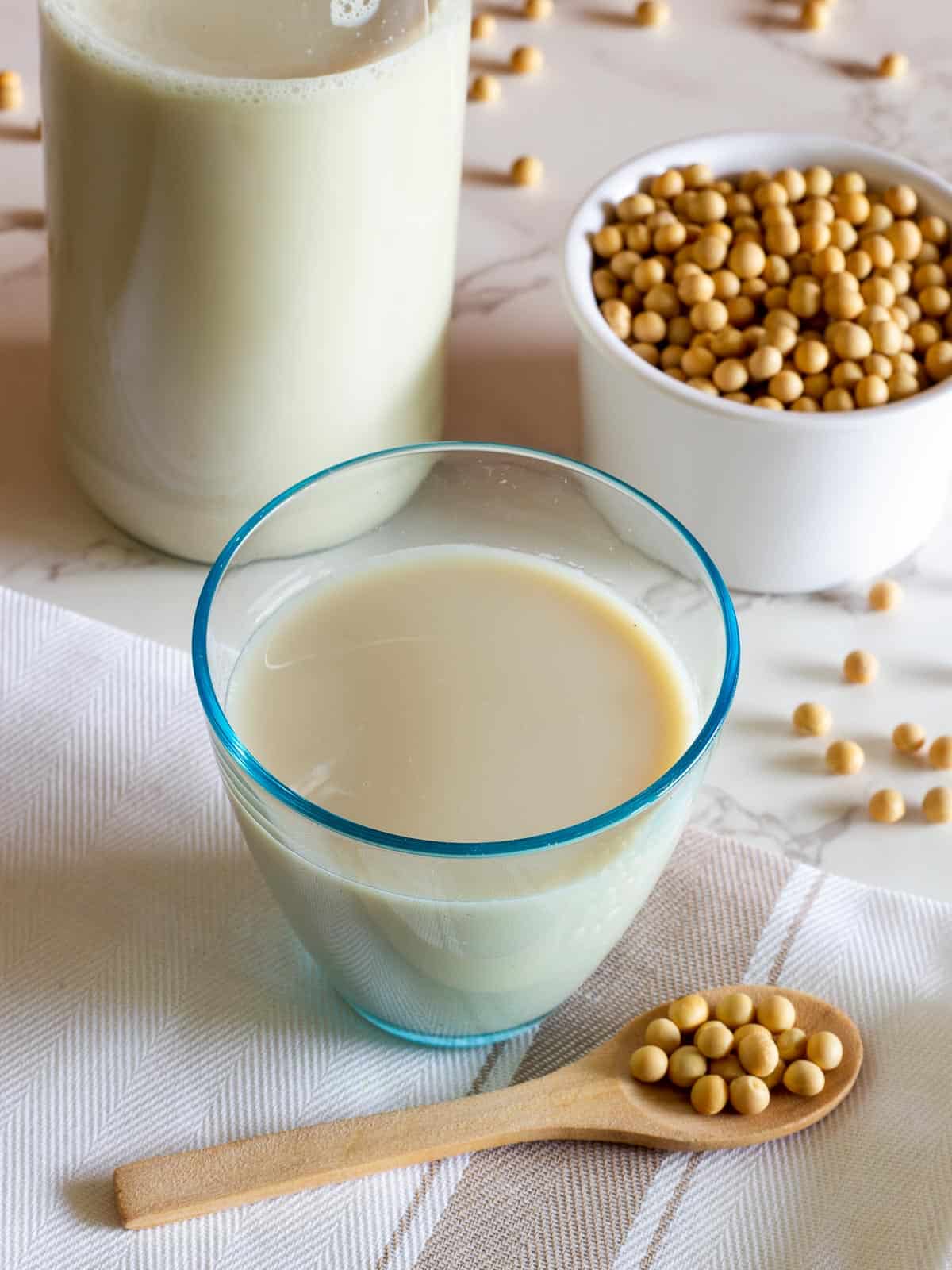 glass of homemade soy milk next to a bottle with milk and dried soybeans spread on a table.