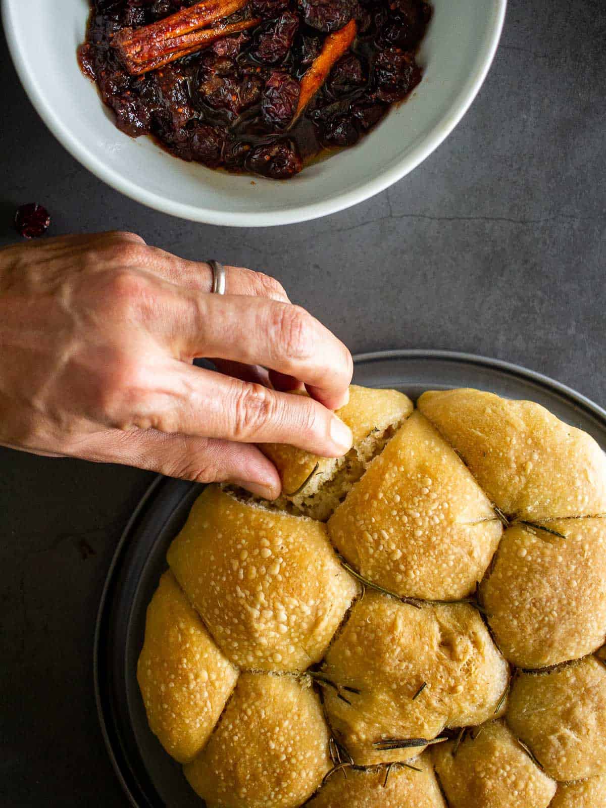 ripping a bread bun from the football shaped baked bread, next to a bowl of cranberry sauce.