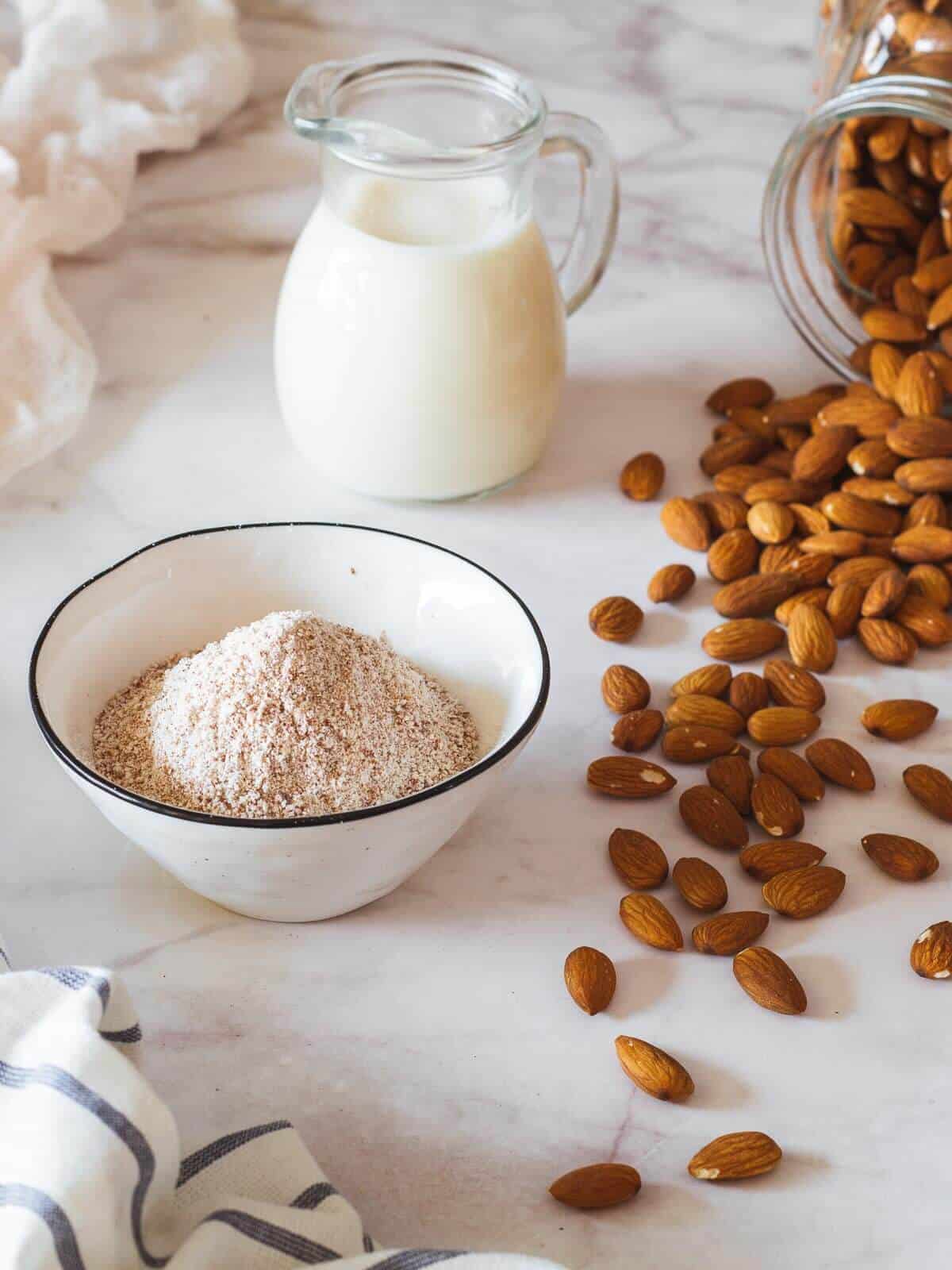 almond meal bowl next to raw almonds, cheesecloth and almond milk vase