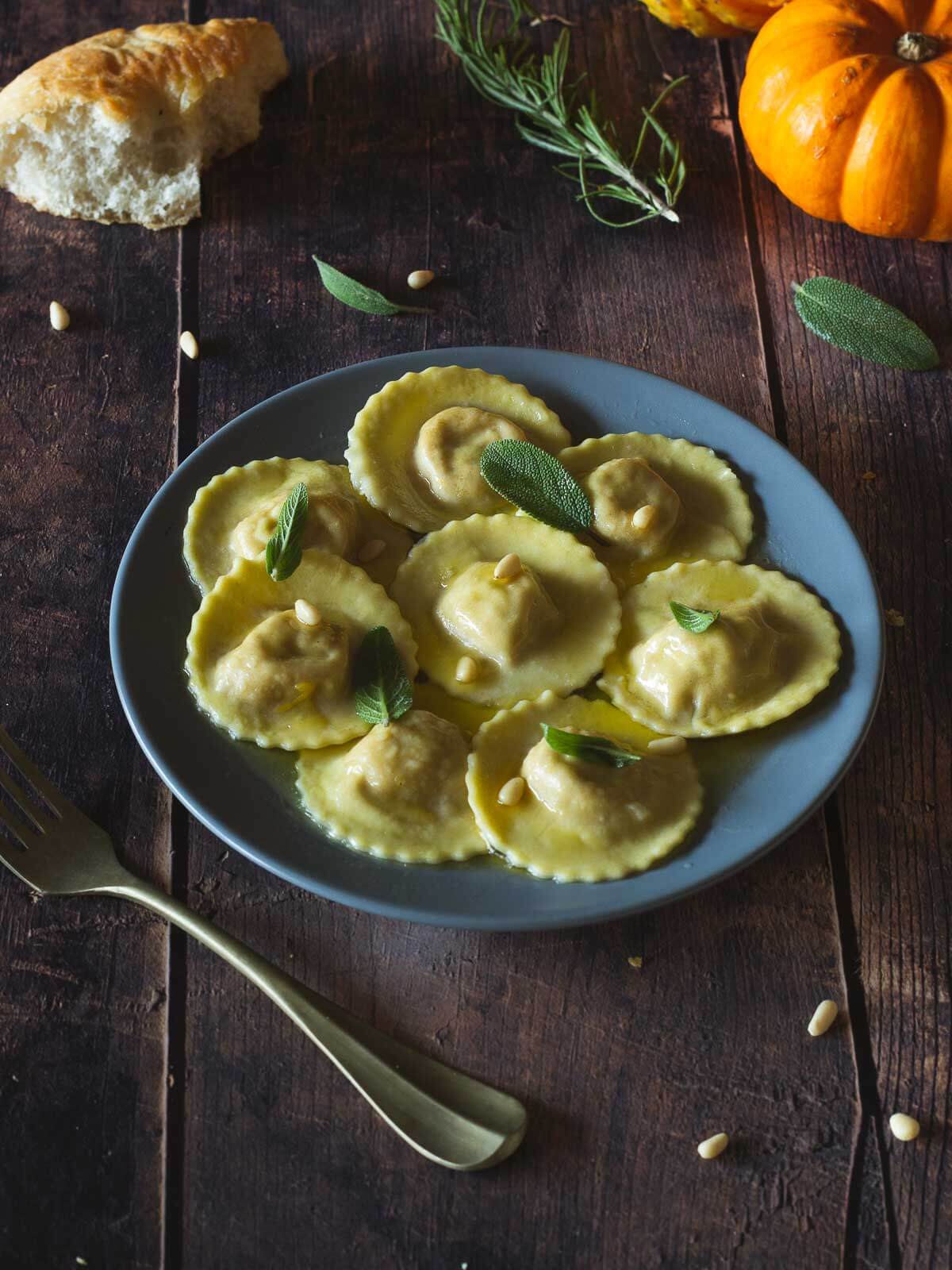 vegan butternut squash ravioli served on a blue flat plate topped with sage butter and pine nuts.
