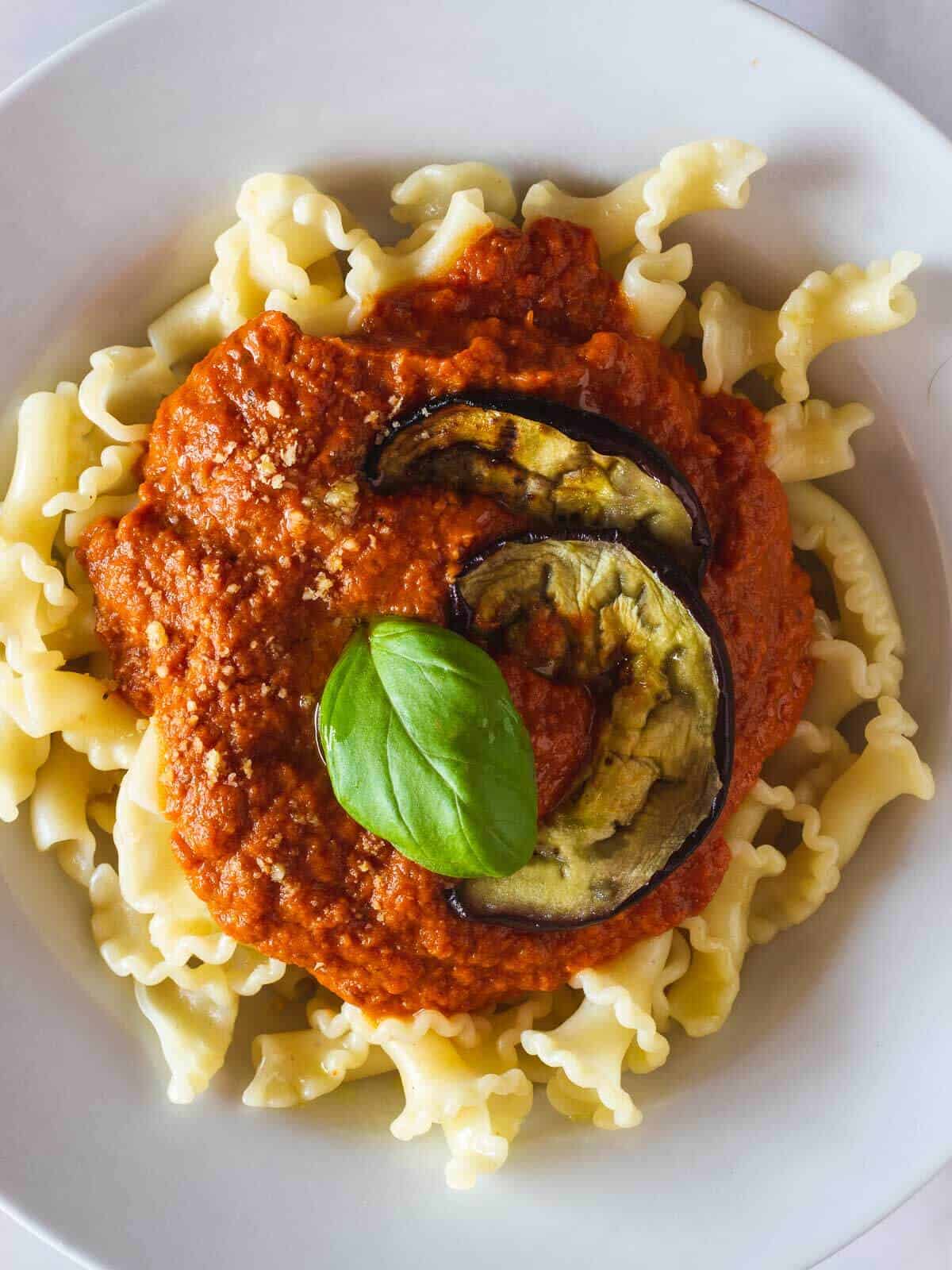 plate served with plain curly pasta and creamy tomato ricotta sauce on top and garnished with grilled eggplant slices and basil leave.
