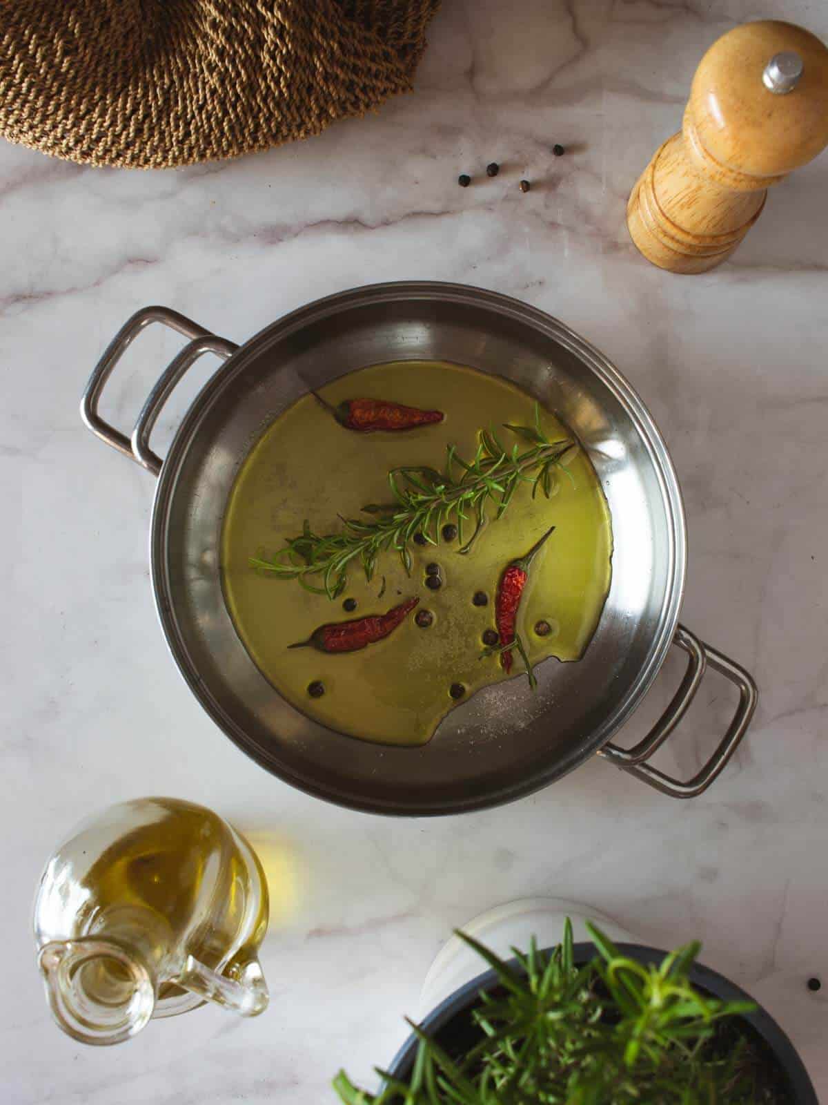 saucepan with olive oil, a rosemary sprig, whole black peppercorns, and pepperoncino chillies.