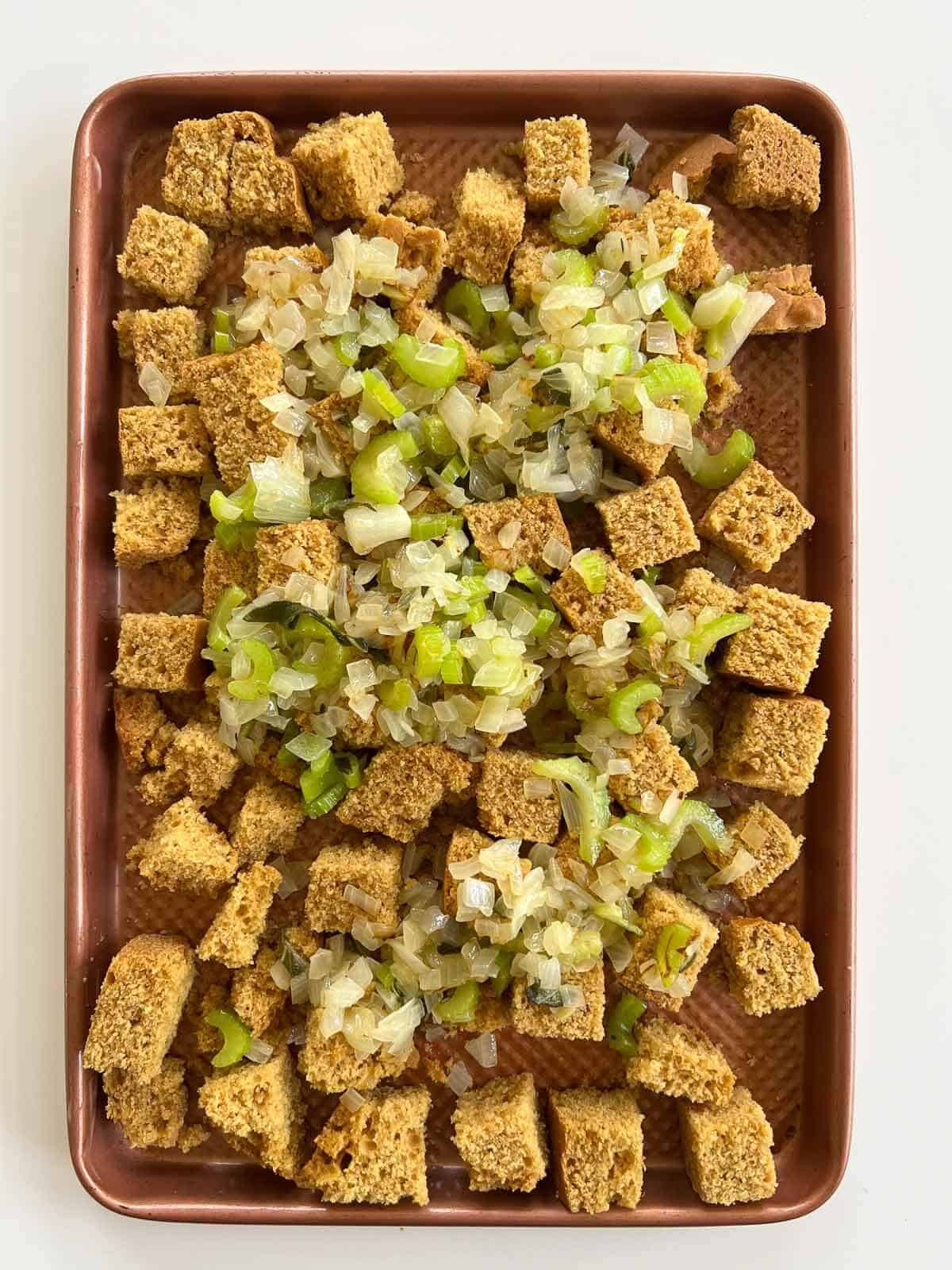 cornbread cubes in baling sheet with cooked onions and celery on top in a baking sheet.