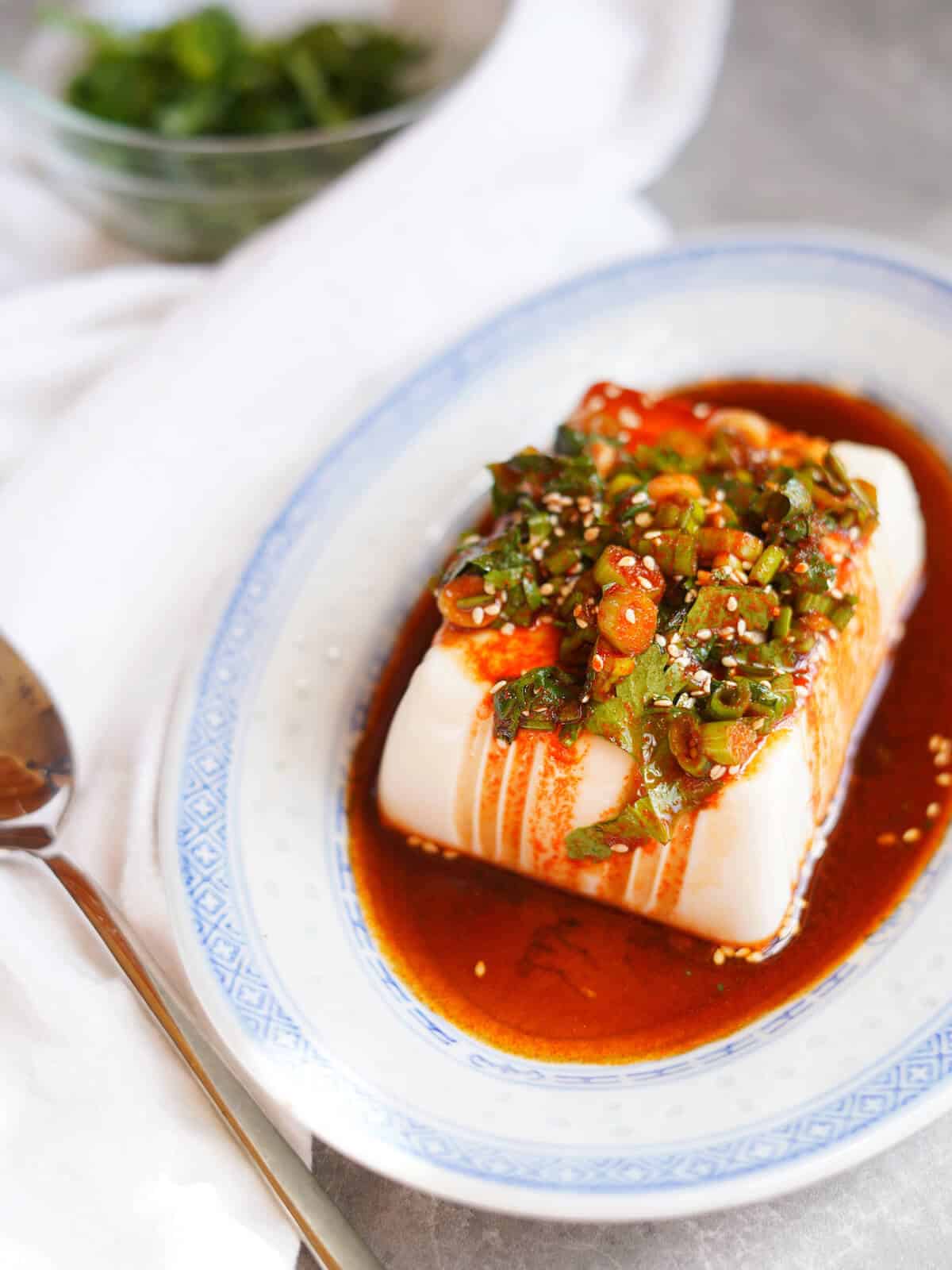 A classic from the soft tofu recipes is this cold tofu served which is served on a plate with a red green onions sauce on top.