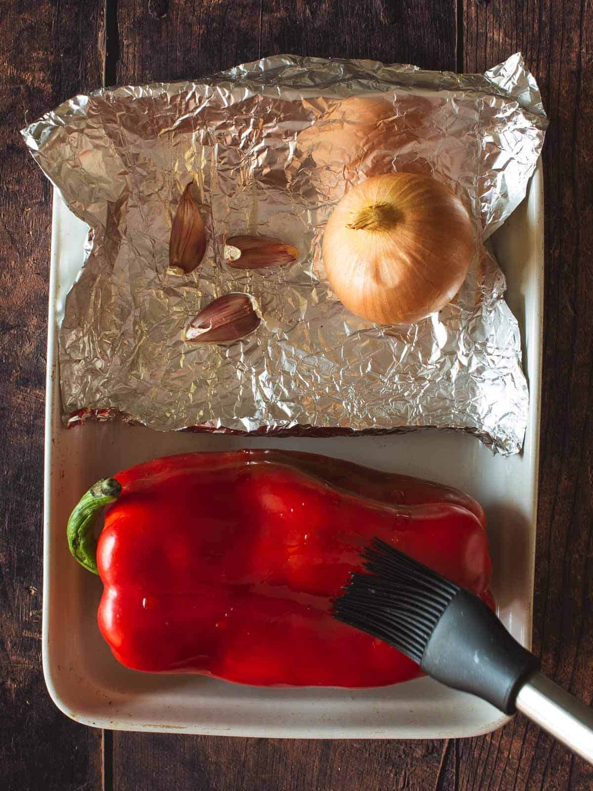 brushing red pepper with olive oil before putting it in the oven, next to an onion and garlic cloves on aluminium foil.
