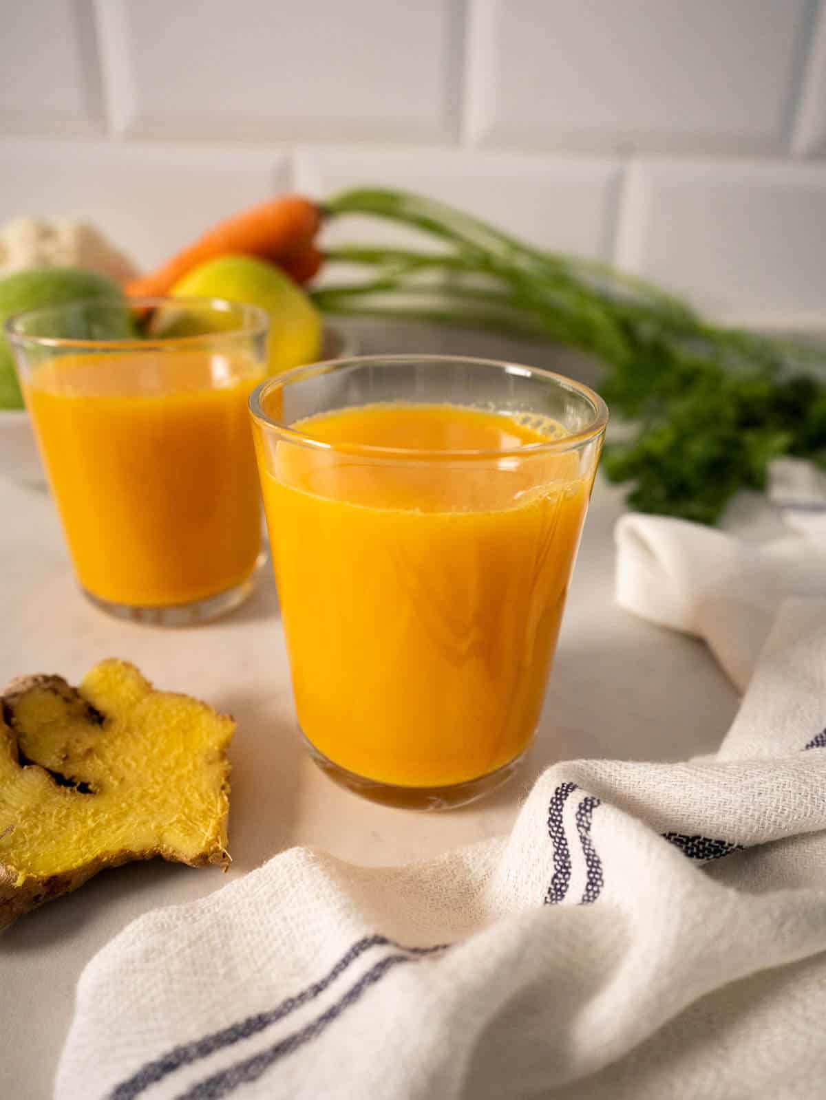 two glasses of carrot ginger turmeric juice with a tablecloth and fresh carrots behind.