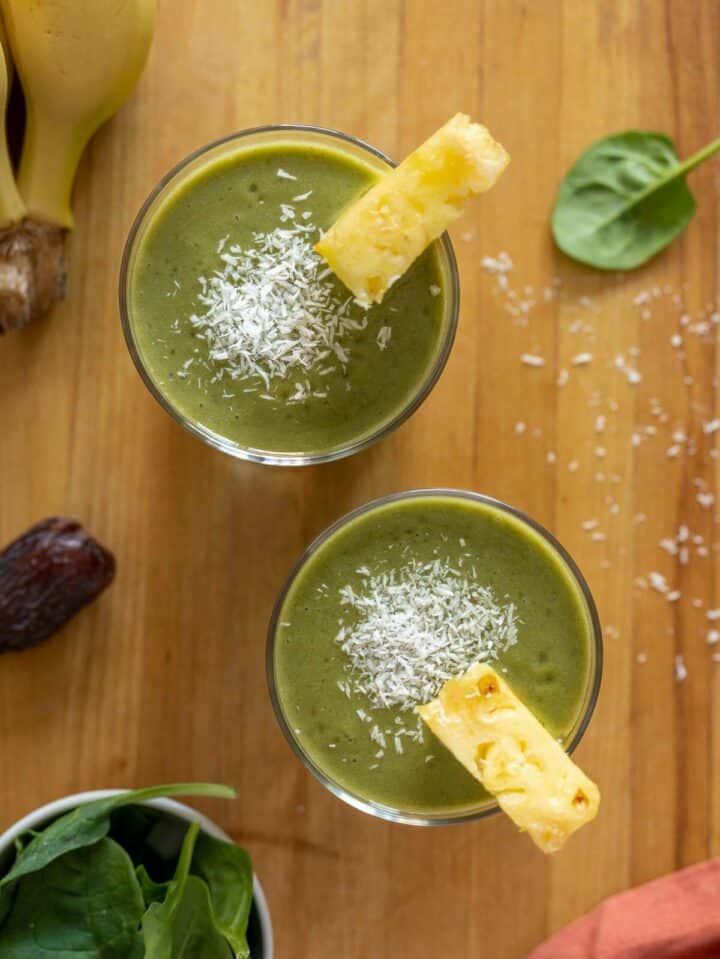 two pineapple banana spinach smoothies on a wooden table seen from above with chunks of fresh pineapple as garnishing.