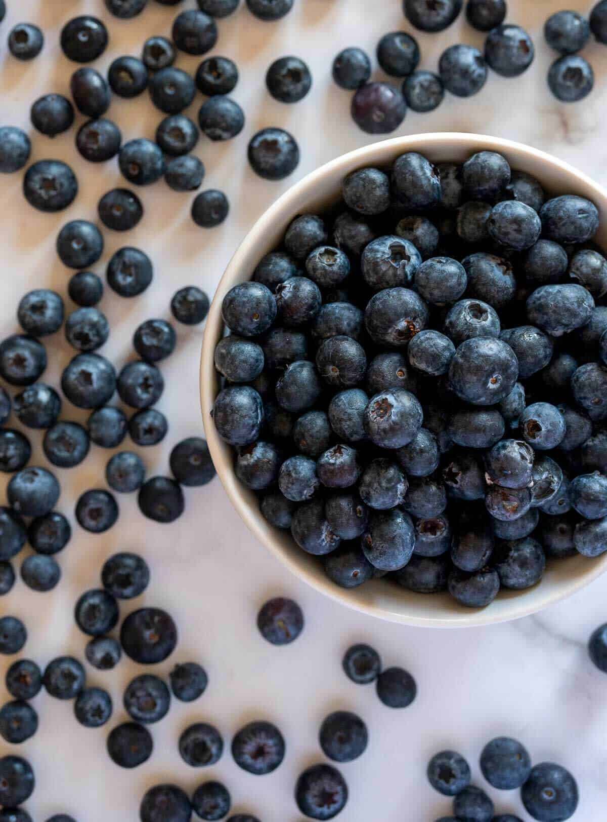 bowl of fresh blueberries and lots of them scattered on a white marble table.