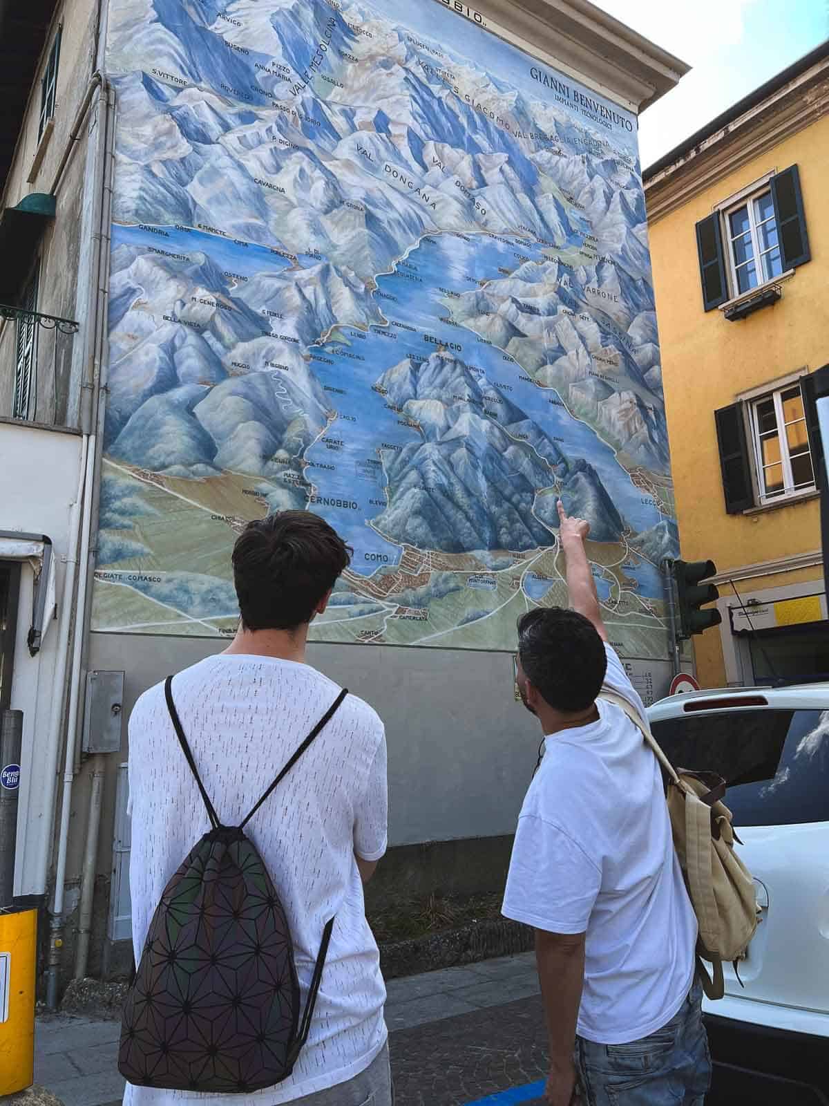 two men deciding where to go watching a painted map on a wall in Cernobbio.