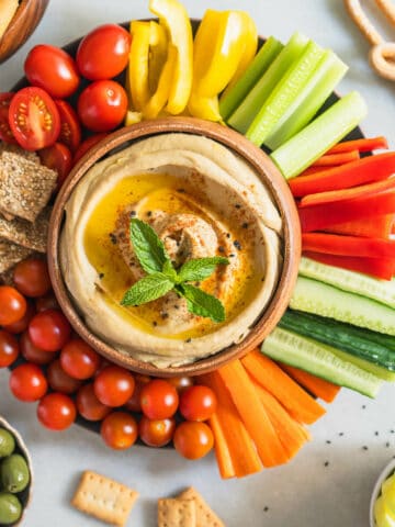 wondering what to eat with hummus? Here is a serving plate with tons of vegetables , cracker and more options with a hummus bowl in the center.