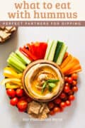 wondering what to eat with hummus? Here is a serving plate with tons of vegetables , cracker and more options with a hummus bowl in the center pin.