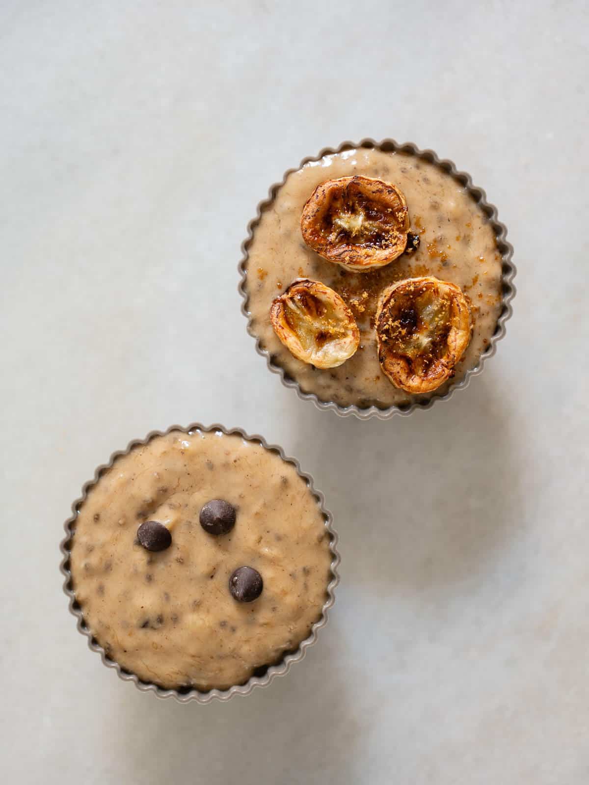 two unbaked muffin tins with batter, one topped with chocolate chips, and others with thinly sliced bananas and brown sugar.