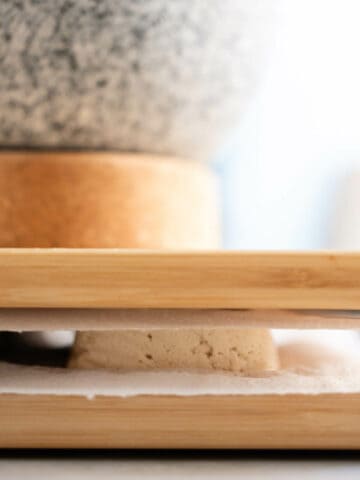 tofu layers between paper towels and chopping boards with something heavy on top to press out the excess moisture.