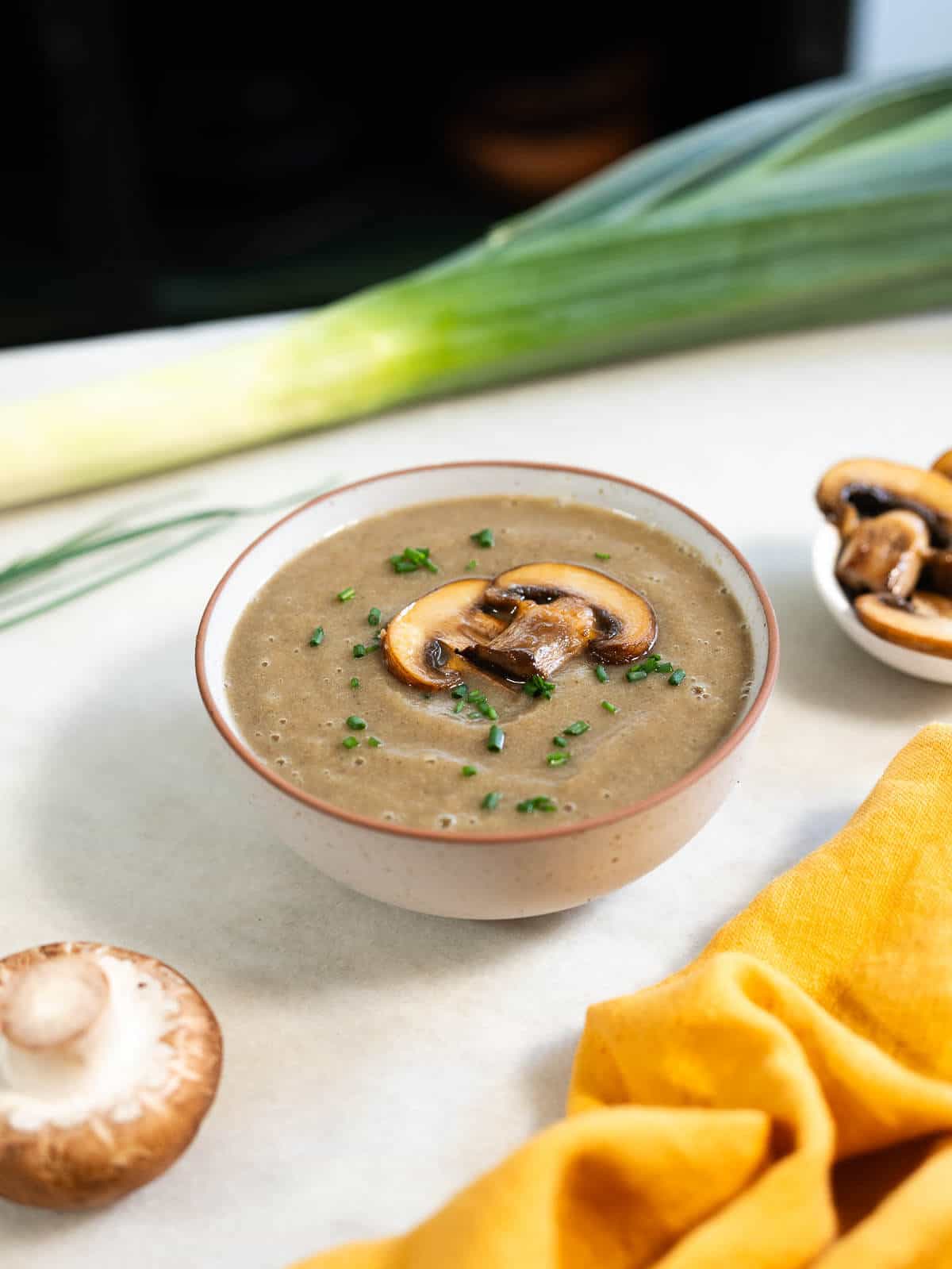 table with a whole leek, small bowl served with mushroom soup without cream garnished with browned mushroom slices and thinly chopped chives.
