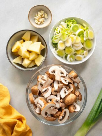 prepared vegetables in bowls: chopped mushrooms, garlic, potatoes, and thinly sliced leeks.