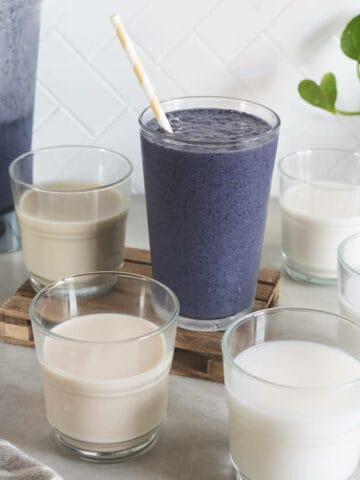 In the center, a vibrant blueberry smoothie in a tall glass with a striped straw stands out against a backdrop of five different glasses of plant milk. The smoothie's rich purple hue suggests a blend of blueberries and plant milk, while the surrounding glasses showcase the variety of milks in shades of white to beige