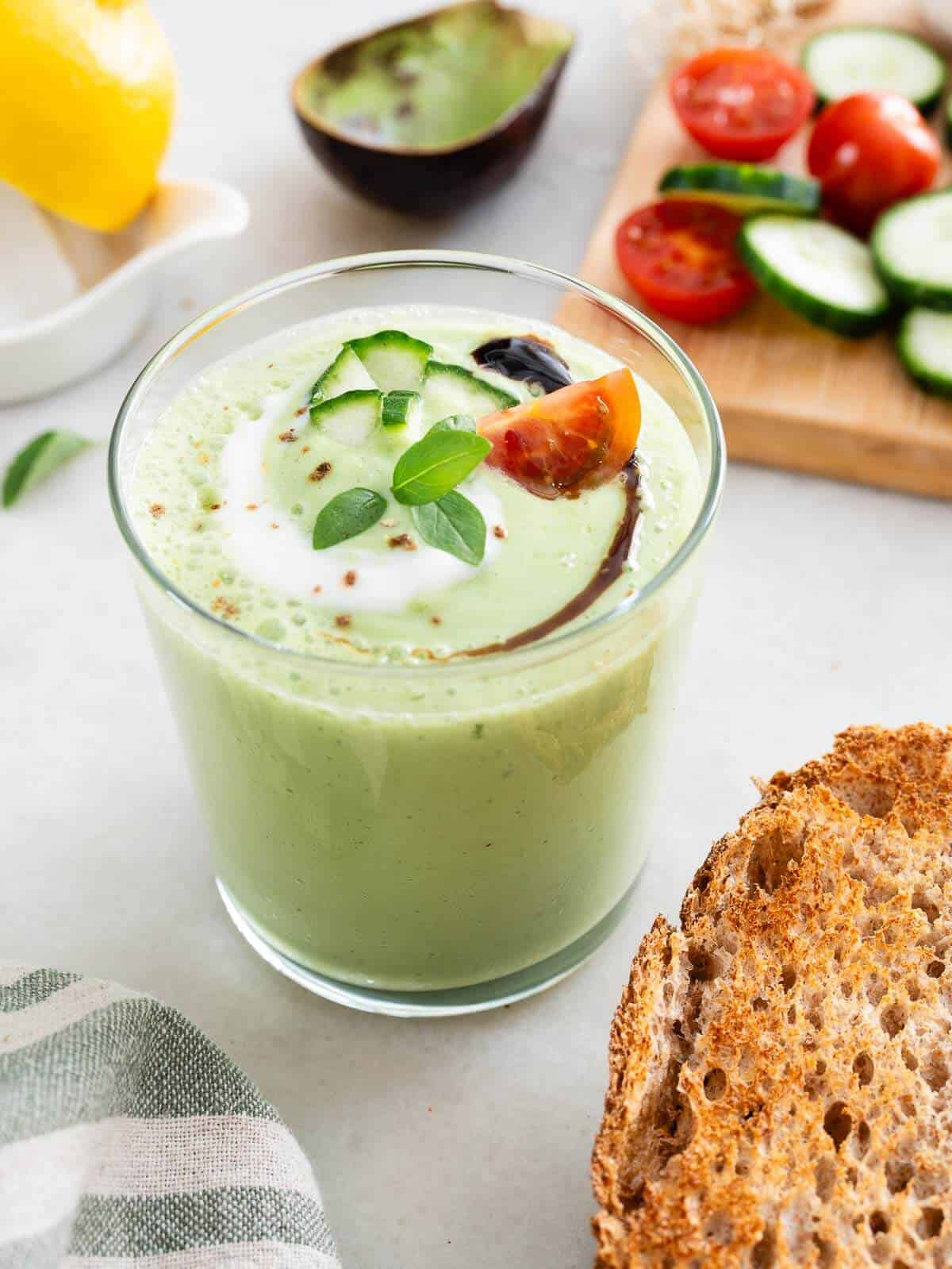 A vibrant glass of creamy green cucumber soup, garnished with a swirl of white yogurt, diced cucumber, a slice of cherry tomato, and fresh basil leaves. In the background, half an avocado, a lemon, sliced cucumbers, cherry tomatoes, and a slice of whole-grain bread rest on a kitchen counter.