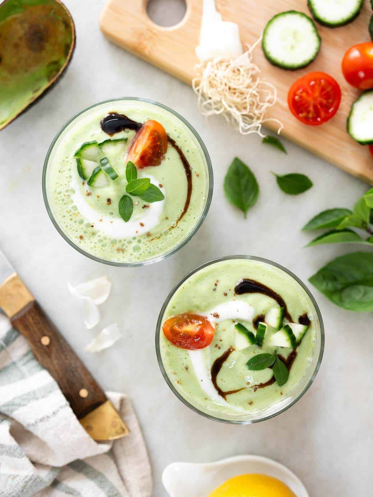 two glasses of chilled basil cucumber soup served with a swirl of balsamic glaze, tomato and cucumber slices, and yogurt.