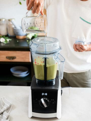 A person pours water from a measuring cup into a high-speed blender containing a vibrant green pasta sauce.