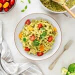 bowl of green high-protein healthy pasta salad garnished with halved cherry tomatoes, broccoli, and fresh basil leaves, accompanied by a bowl of cherry tomatoes.