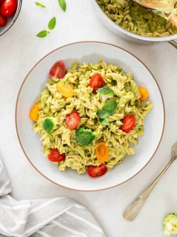 bowl of green high-protein healthy pasta salad garnished with halved cherry tomatoes, broccoli, and fresh basil leaves, accompanied by a bowl of cherry tomatoes.