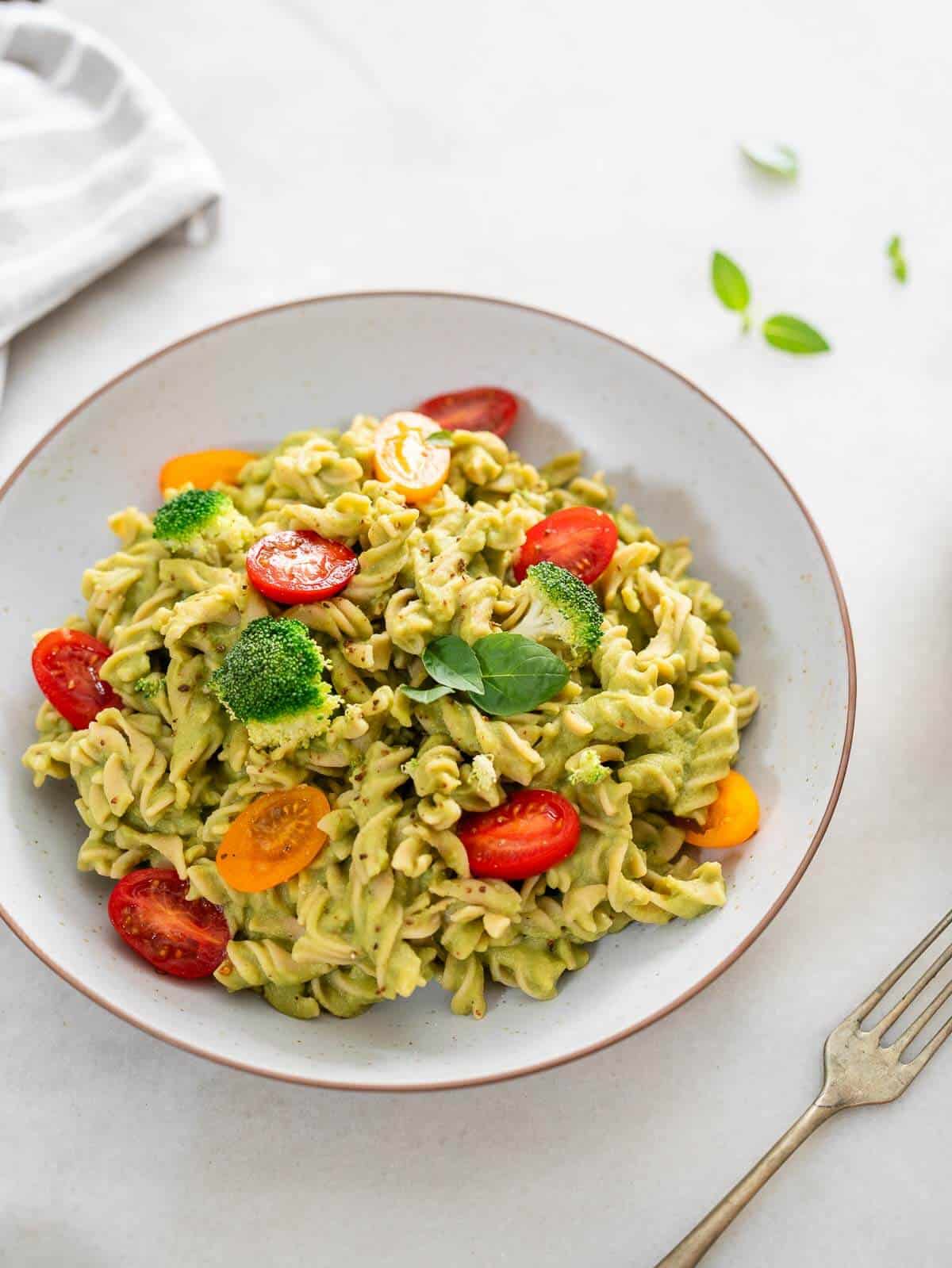 A bowl of creamy green healthy pasta salad topped with cherry tomatoes, broccoli florets, and fresh basil,