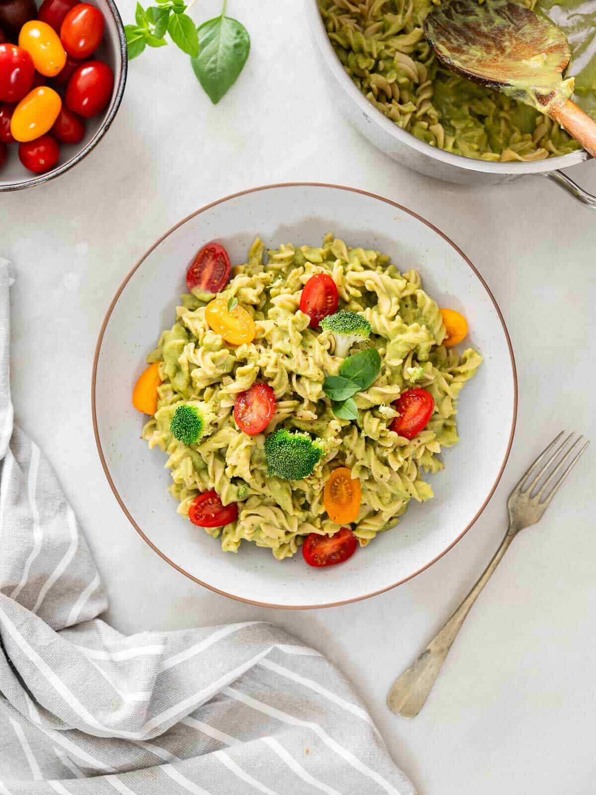 bowl of green high-protein healthy pasta salad garnished with halved cherry tomatoes, broccoli, and fresh basil leaves, accompanied by a bowl of cherry tomatoes and a kitchen towel