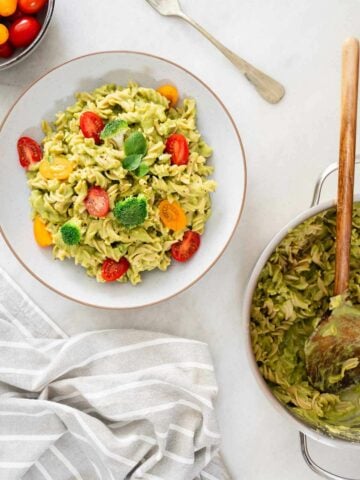 A bowl of green pasta salad is being served with a wooden spoon, showcasing the creamy texture and mix of vegetables, with a pan of more pasta salad visible in the background.