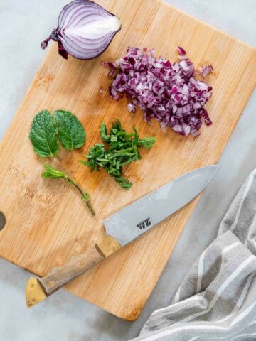 Fresh red onion and spearmint being finely chopped on a wooden cutting board.
