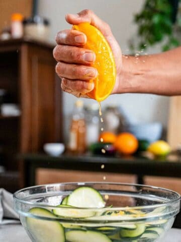 squeezing a fresh orange on top of a bowl with sliced zucchini.