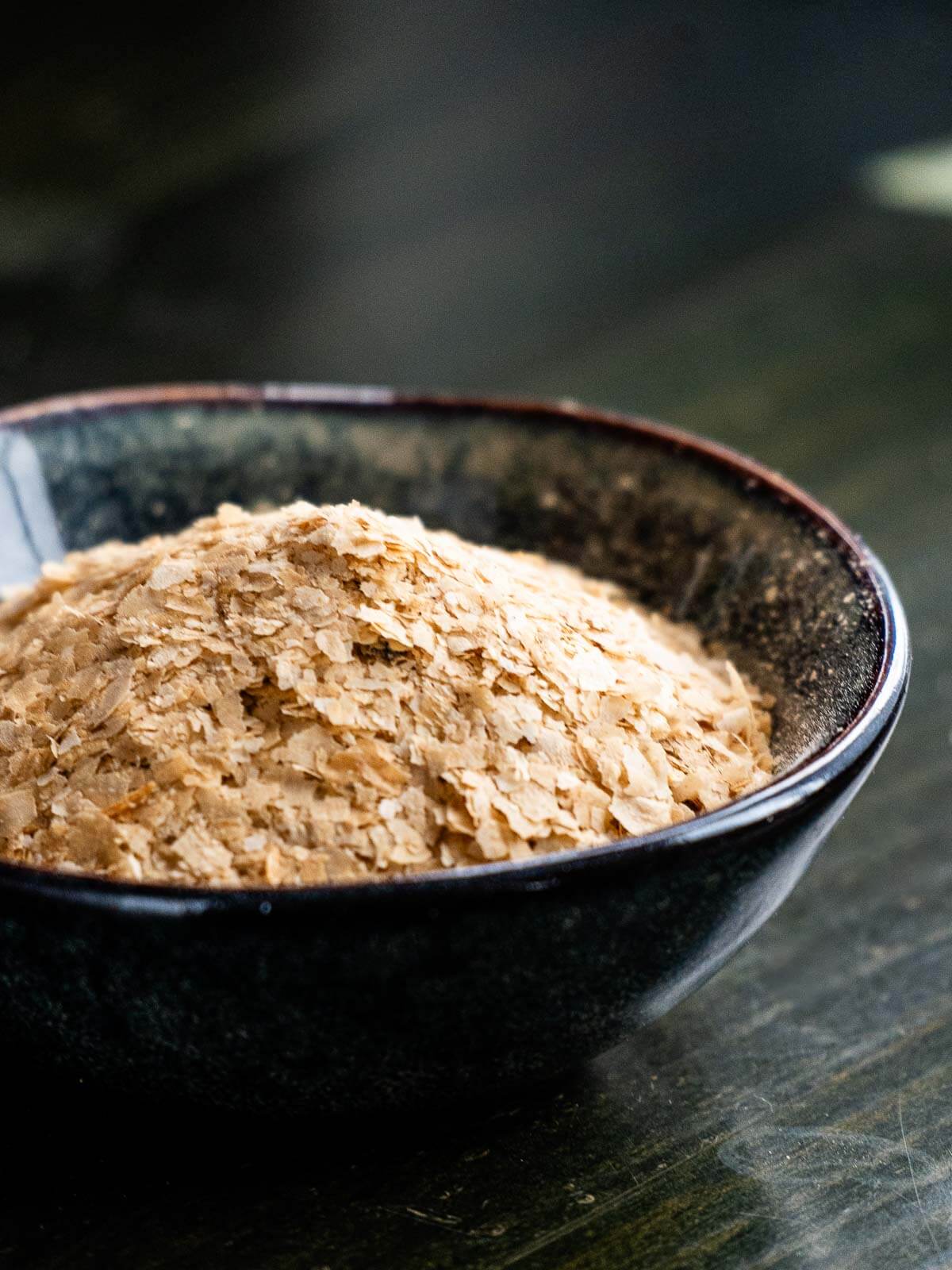 A small black bowl filled with nutritional yeast flakes placed on a dark surface, emphasizing the rich, golden color of the yeast against the contrasting background.