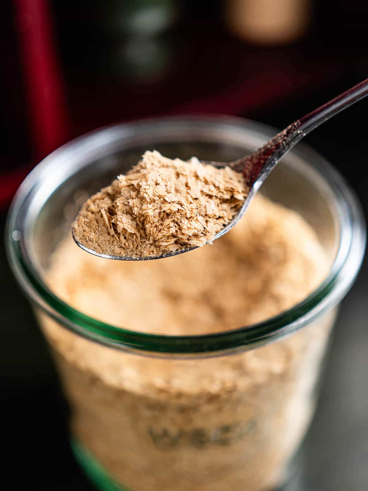 A spoonful of nutritional yeast flakes held above a glass jar filled with more of the flakes, showcasing a close-up view that highlights the texture of the yeast.