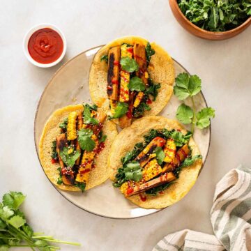 Three yellow lentil wraps served in a plate with a pan-fried seitan and corn strips filling, topped with sriracha sauce and coriander leaves.